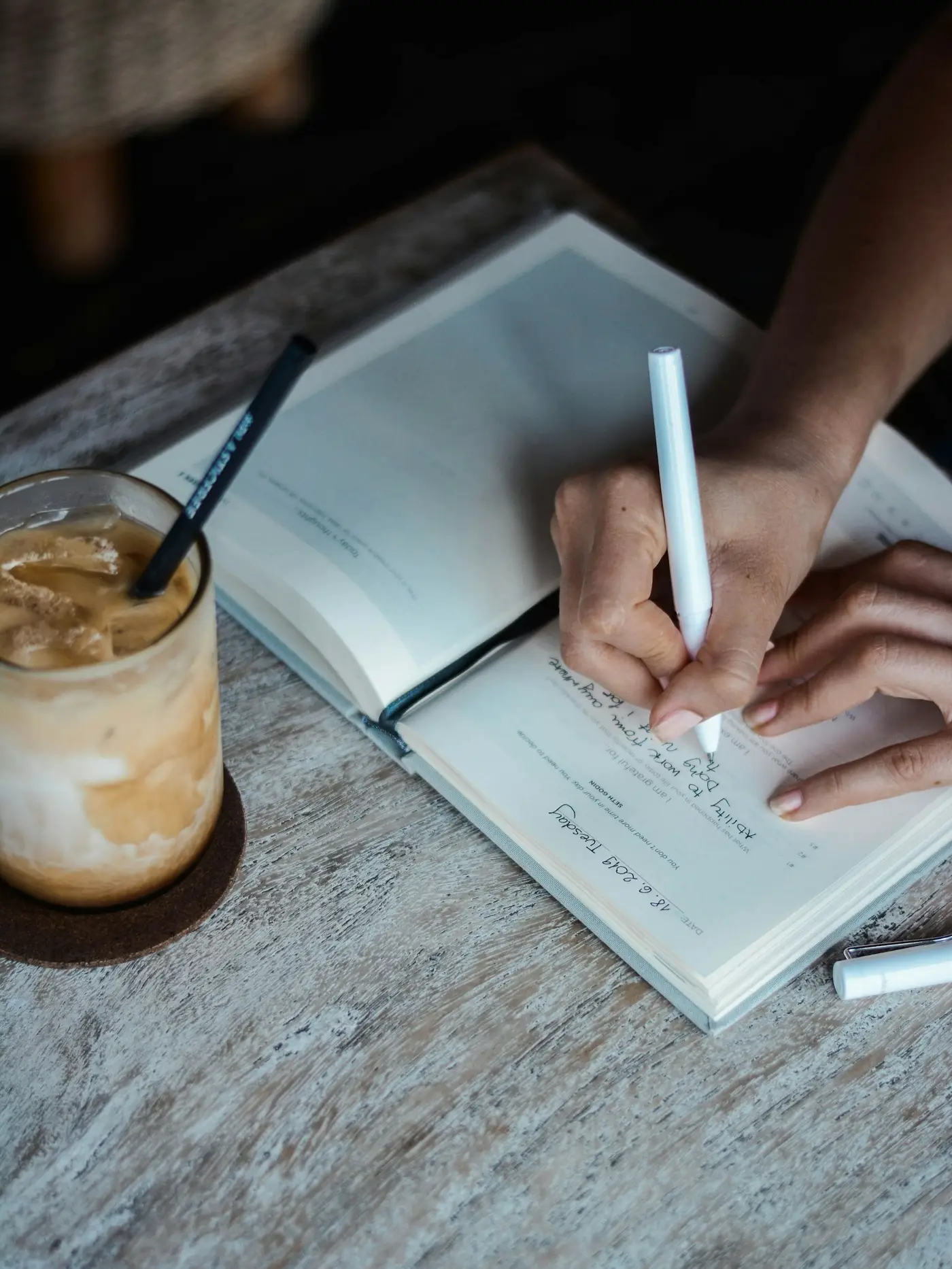 Hand schreibt in ein Notizbuch, Eiskaffee daneben, Holztisch