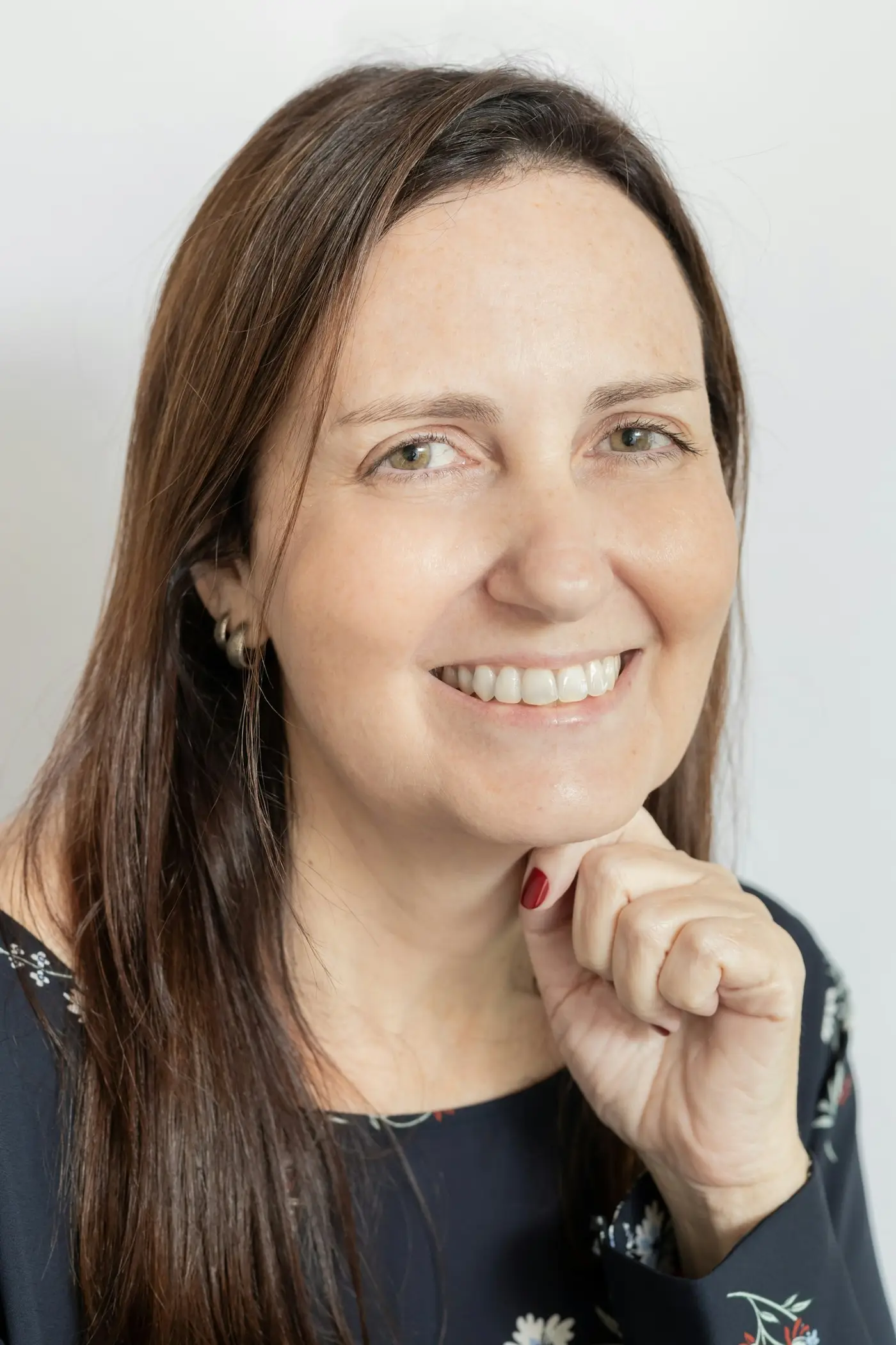 Frau um 45, lange braune Haare, breites Lächeln, Blick in die Kamera, Hand am Kinn, heller grauer Hintergrund