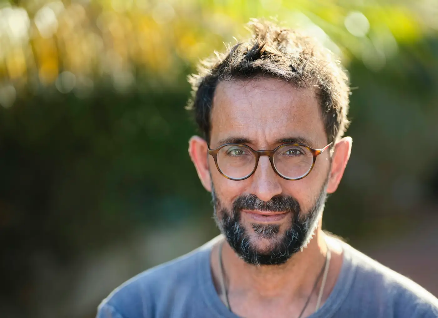 Mann um 45, Rundbrille, grauer Bart, blaues T-Shirt, leichtes Lächeln, warmes Gegenlicht draußen