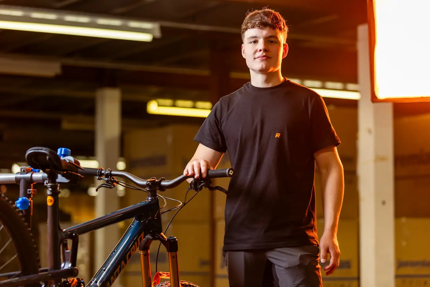 Junger Mitarbeiter steht mit Mountainbike im Laden/Lager, Blick in die Kamera, warmes Licht, keine Masken